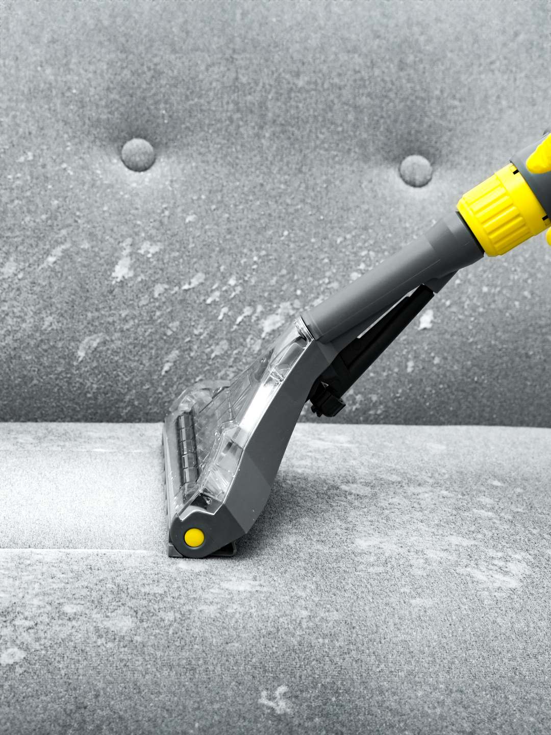 A close-up of a vacuum cleaner attachment being used on a gray upholstered couch, with visible dust and debris being suctioned from the fabric.