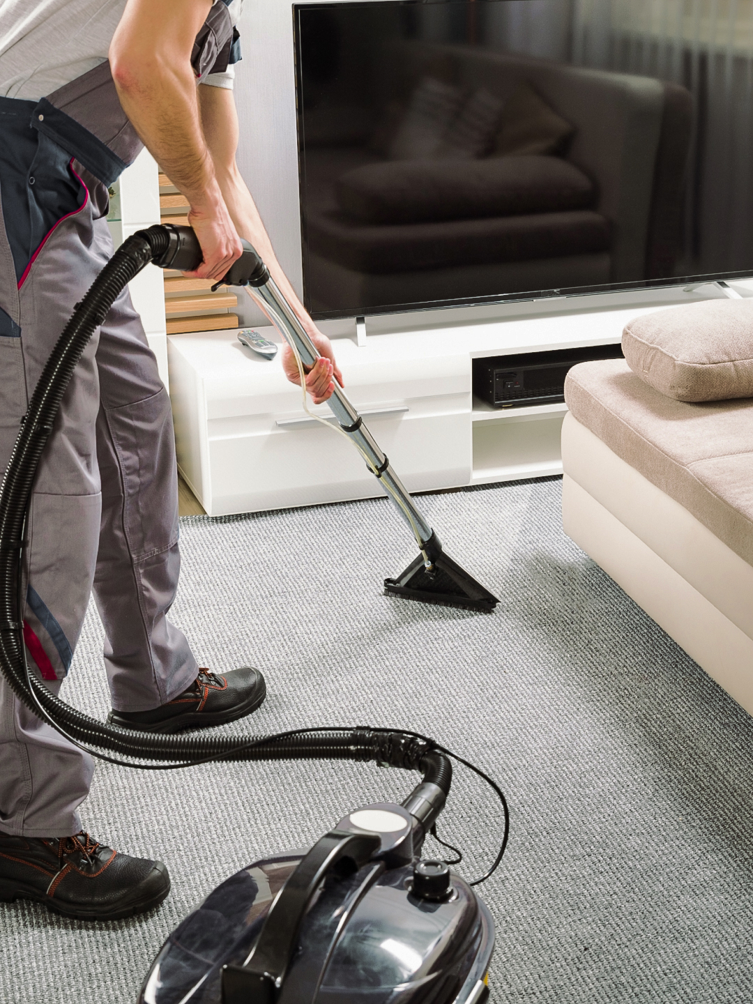 A person in work attire vacuuming a carpeted living room with a vacuum cleaner, featuring a large television in the background and a light-colored sofa.